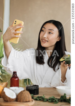 A young Asian woman in a white bathrobe takes a selfie with her phone while holding avocado and skincare products, preparing for a beauty routine A young Asian woman in a white bathrobe takes a selfie with her phone while holding avocado and skincare products, preparing for a beauty routine 135535969
