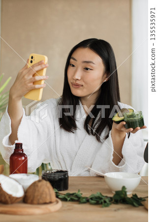 A young Asian woman in a white bathrobe films her natural skincare routine with avocado and coconut ingredients on her phone 135535971