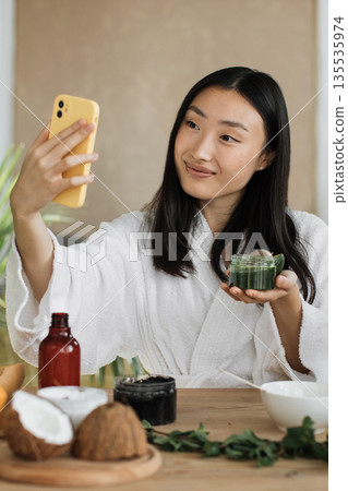 A young Asian woman in a white bathrobe holds a jar of green skincare product and a smartphone, preparing to take a selfie while surrounded by natural ingredients like coconut and herbs 135535974