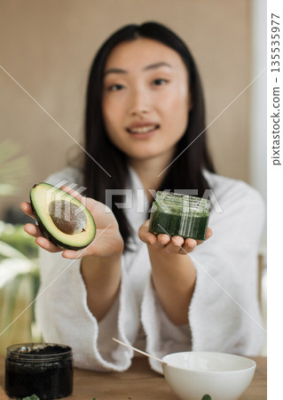 A young woman in a white robe holds a halved avocado in one hand and a jar of green cosmetic mask in the other, suggesting natural skincare A young woman in a white robe holds a halved avocado in one hand and a jar of green cosmetic mask in the other, suggesting natural skincare 135535977
