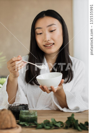 A young woman in a white robe holds a spoon with cream over a bowl, preparing for a skincare routine A young woman in a white robe holds a spoon with cream over a bowl, preparing for a skincare routine 135535991