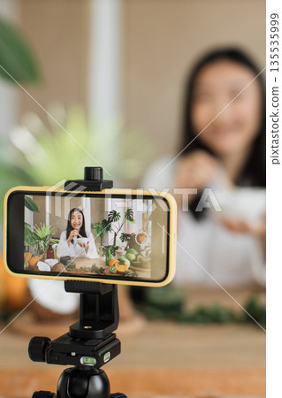 A young woman films herself preparing a healthy meal for her online audience, surrounded by fresh ingredients and plants A young woman films herself preparing a healthy meal for her online audience, surrounded by fresh ingredients and plants 135535999