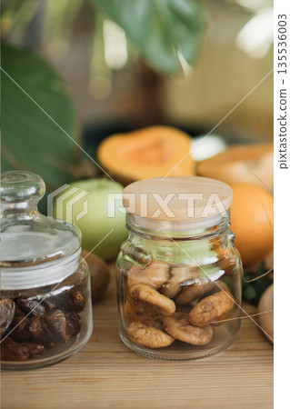 Two glass jars filled with dried fruits, dates and figs, sit on a wooden surface with fresh fruits in the background Two glass jars filled with dried fruits, dates and figs, sit on a wooden surface with fresh fruits in the background 135536003