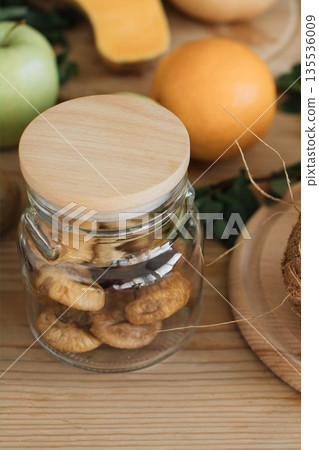 A glass jar filled with dried figs sits on a wooden table surrounded by fresh fruits and vegetables 135536009