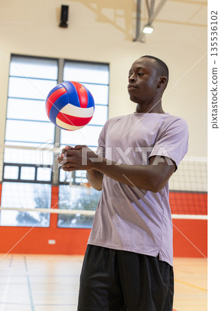 African american male athlete spiking red white, blue volleyball over net inside gymnasium court 135536102
