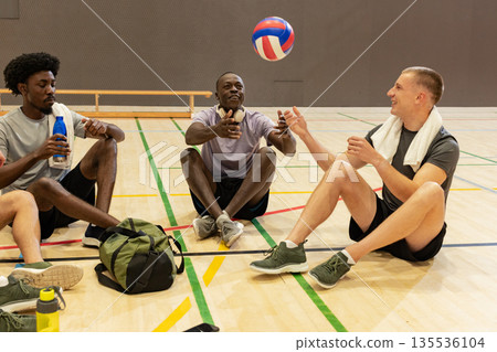 Diverse male partners sitting on sports hall floor tossing red white blue volleyball with towel 135536104