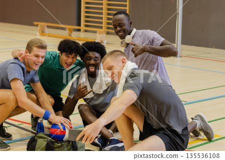 Diverse male teammates crouching on gym court checking smartphone near volleyball and duffel bag Diverse male teammates crouching on gym court checking smartphone near volleyball and duffel bag 135536108