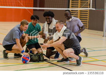 Diverse male volleyball players kneeling in gym under net laughing around smartphone, volleyball 135536110