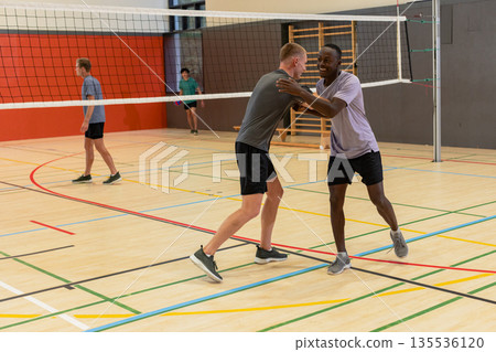 Diverse male partners exchanging high five at net inside gymnasium during volleyball match 135536120