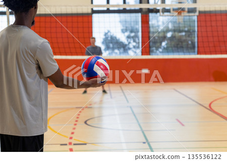 Diverse male volleyball teammates training in gym, holding volleyball next to net and hoop 135536122