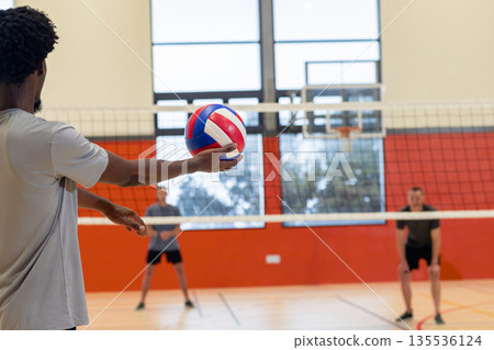 Diverse teammates serving, receiving volleyball on polished wooden court with net, copy space 135536124