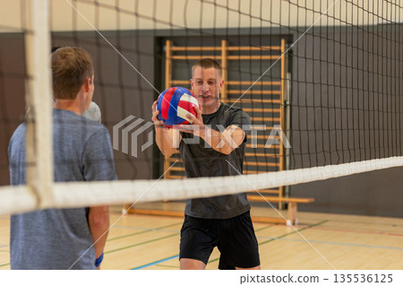 Diverse male teammates practicing volleyball drill in school gym with red white blue ball 135536125