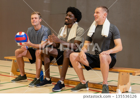 Diverse male teammates sitting on gym bench in sportswear holding volleyball, towels Diverse male teammates sitting on gym bench in sportswear holding volleyball, towels 135536126