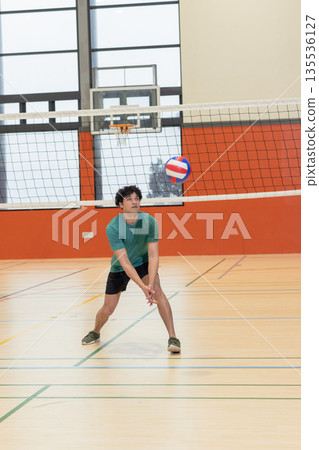 Teenage male volleyball athlete in shirt passing volleyball in gym under net with basketball hoop 135536127