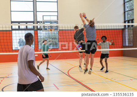 Diverse male teammates jumping to block red, white and blue volleyball at gym net on wooden court 135536135