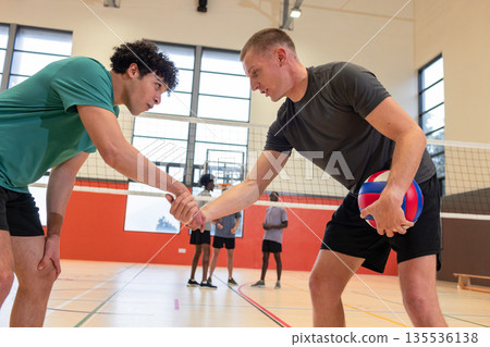 Male volleyball players shaking hands at net in school gym near bench, ball, court markings Male volleyball players shaking hands at net in school gym near bench, ball, court markings 135536138