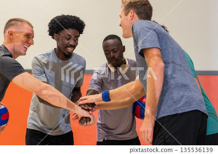 Diverse male teammates stacking hands in sports hall holding volleyballs, wearing headphones Diverse male teammates stacking hands in sports hall holding volleyballs, wearing headphones 135536139