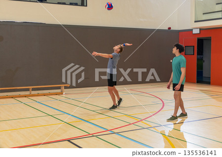 Two male volleyball players serving ball, watching in gym on court with bench, boundary lines Two male volleyball players serving ball, watching in gym on court with bench, boundary lines 135536141