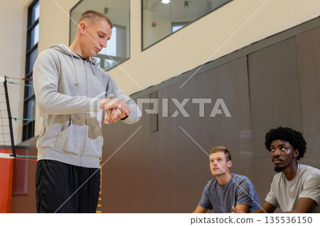 Diverse volleyball coach checking wristwatch and players kneeling beside net on floor lines 135536150