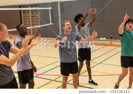 Diverse male volleyball team clapping, raising arms on gym court under net with water bottle Diverse male volleyball team clapping, raising arms on gym court under net with water bottle 135536151