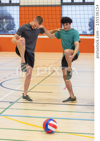 Diverse male partners stretching knees to chest on gymnasium court with volleyball and net 135536154