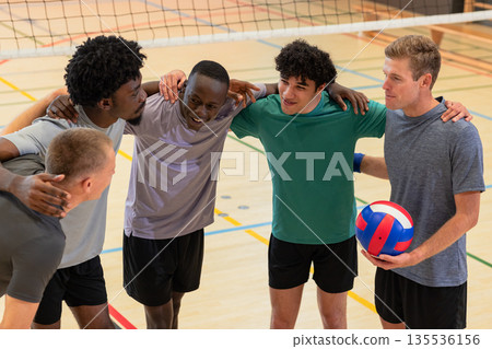 Diverse male partners huddling on gym volleyball court holding volleyball beneath net posts 135536156