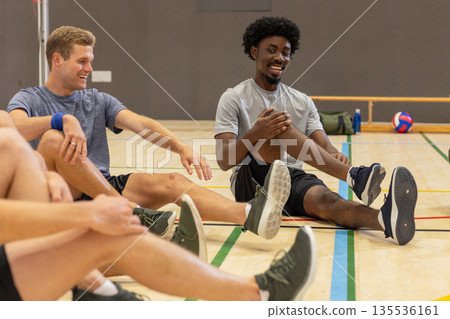 Diverse male teammates in sportswear sitting on floor stretching and chatting next to volleyball Diverse male teammates in sportswear sitting on floor stretching and chatting next to volleyball 135536161