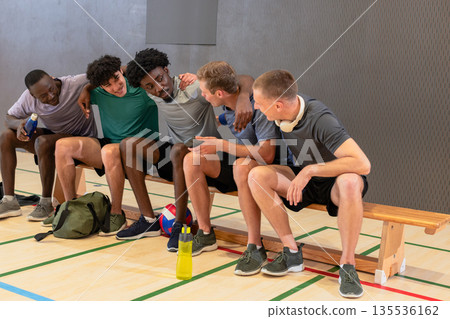Diverse male partners sitting on bench at gym, chatting after volleyball game with water bottles Diverse male partners sitting on bench at gym, chatting after volleyball game with water bottles 135536162