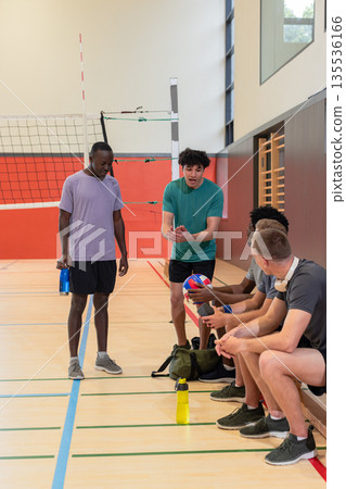 Diverse male teammates gathering beside volleyball net in gym holding water bottles, volleyballs 135536166