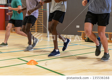 Diverse male partners performing footwork drill around orange cone in gym under volleyball net Diverse male partners performing footwork drill around orange cone in gym under volleyball net 135536168