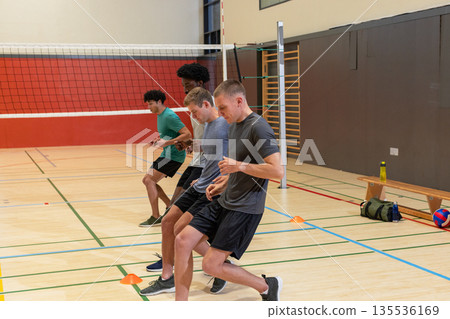 Diverse male players practicing footwork drills at gym using orange cones beside volleyball net 135536169