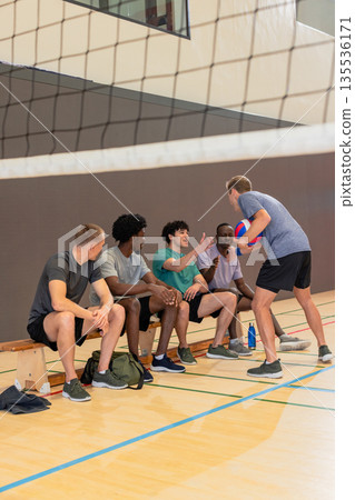 Diverse male volleyball partners sitting on bench and standing holding volleyball in school gym Diverse male volleyball partners sitting on bench and standing holding volleyball in school gym 135536171