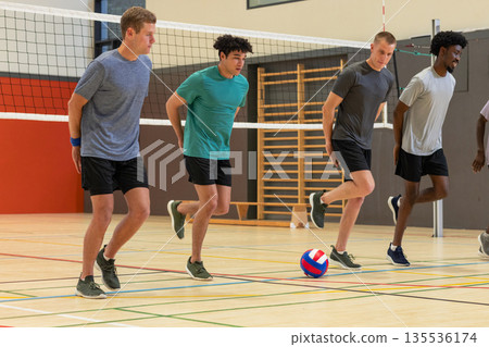 Diverse male volleyball partners performing knee-raise warm-up drill inside gymnasium by net Diverse male volleyball partners performing knee-raise warm-up drill inside gymnasium by net 135536174