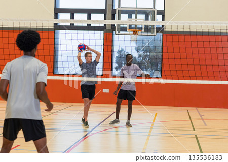 Diverse male partners serving volleyball across net on volleyball court with basketball hoop Diverse male partners serving volleyball across net on volleyball court with basketball hoop 135536183
