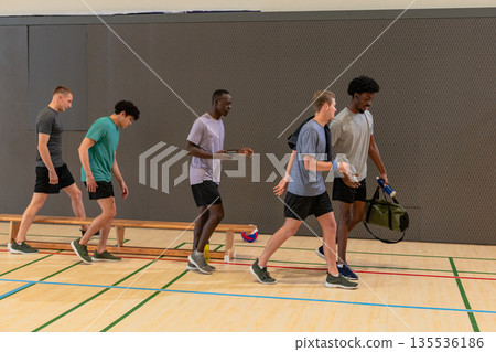 Male friends walking in gym passing bench with volleyball, carrying duffel bag, tennis racket 135536186