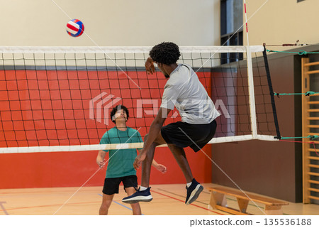Diverse male friends rallying volleyball over net at school gym with bench, climbing bars 135536188