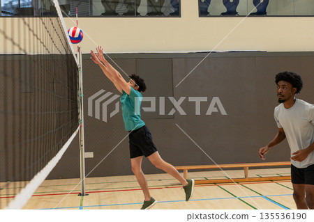 Male volleyball players in teal, grey shirts blocking red-white-blue volleyball at net in gym 135536190