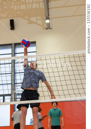 Diverse male teammates spiking volleyball at net in gym with red panels and overhead light 135536192