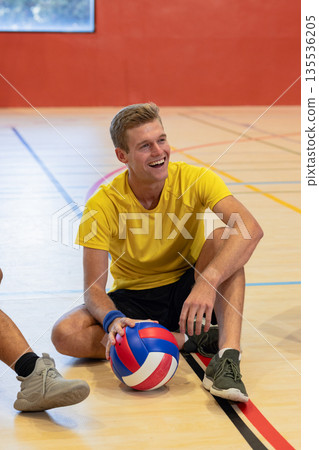 Man sitting on gymnasium court holding volleyball wearing yellow shirt, wristband 135536205