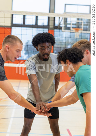 Diverse male teammates in gear huddling, stacking hands under volleyball net on court with hoop Diverse male teammates in gear huddling, stacking hands under volleyball net on court with hoop 135536210
