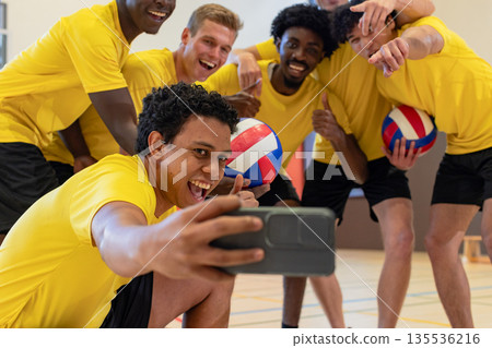 Diverse male volleyball teammates posing for group selfie on court with smartphone, volleyballs 135536216