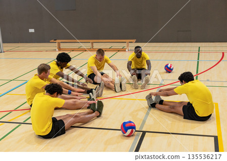 Diverse male volleyball team stretching legs forward on lined court next to volleyballs Diverse male volleyball team stretching legs forward on lined court next to volleyballs 135536217