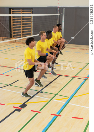 Diverse male partners performing synchronized knee lifts in sports hall with volleyball net posts Diverse male partners performing synchronized knee lifts in sports hall with volleyball net posts 135536221