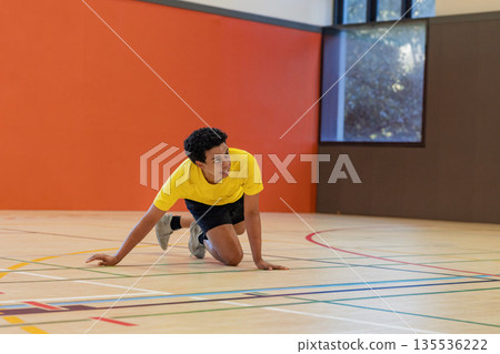 Teenage african american male kneeling on gym court bracing on colored floor lines, sneakers Teenage african american male kneeling on gym court bracing on colored floor lines, sneakers 135536222