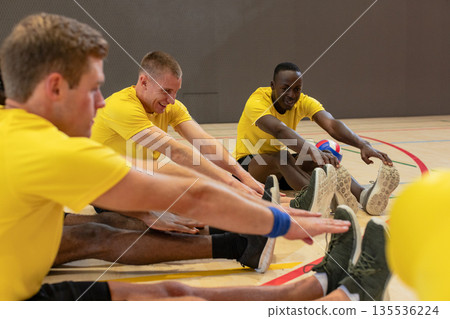 Male volleyball teammates in yellow shirts stretching legs on gym court holding volleyball Male volleyball teammates in yellow shirts stretching legs on gym court holding volleyball 135536224