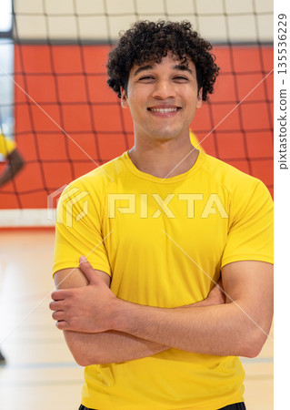 Male player standing with arms crossed before volleyball net while smiling inside gym court 135536229