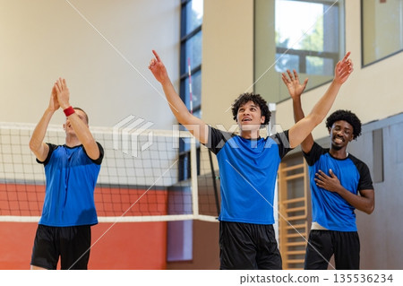 Diverse male volleyball athletes celebrating point in gym at volleyball net in blue black jerseys 135536234
