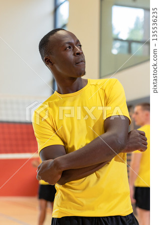 African american male player crossing arms by volleyball net in gym wearing yellow jersey shorts African american male player crossing arms by volleyball net in gym wearing yellow jersey shorts 135536235