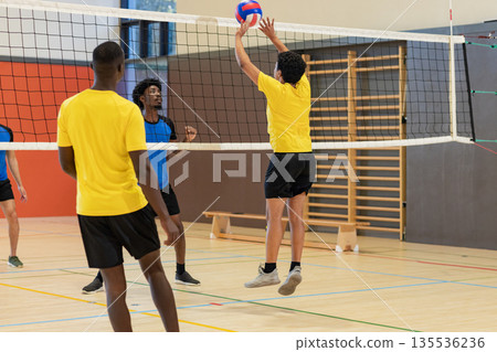 Diverse male volleyball players leaping and blocking on gym court with net, red-white-blue ball 135536236