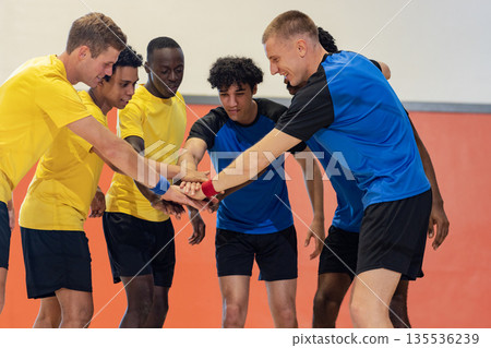 Diverse male athletes huddling placing hands at center in gymnasium in yellow and blue jerseys 135536239
